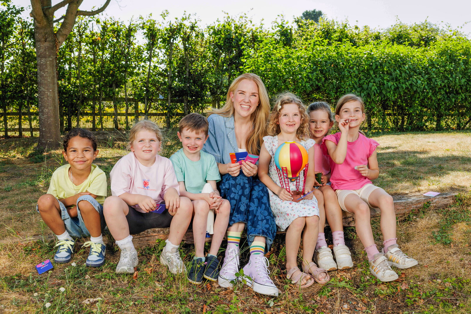 Maddie Moate sitting on a log with a group of smiling children holding prompt cards